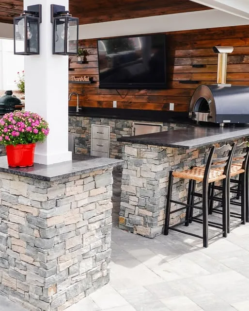 Covered outdoor kitchen with a stone bar, stools, built-in grill and pizza oven